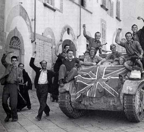 immagine storica in bianco e nero legata all'esultanza collettiva per la liberazione della città di Arezzo durante la Seconda Guerra Mondiale. Al centro e sulla destra domina un mezzo corazzato cingolato, sul cui fronte è chiaramente visibile e stesa una grande bandiera del Regno Unito. Sopra e a bordo del mezzo militare ci sono diversi soldati in divisa.
